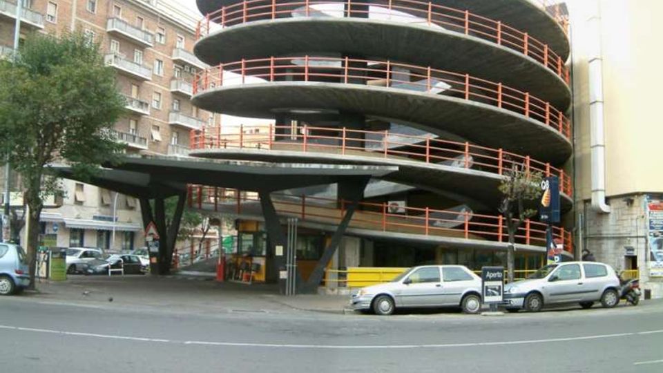 Parcheggio multipiano a rampa elicoidale con grandi solai circolari in cemento e ringhiere arancioni, affacciato su una strada urbana. Alla base si vede un ingresso coperto con pilastri inclinati, con edifici residenziali e auto parcheggiate intorno.