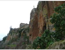 Borgo con edifici in pietra arroccati sul bordo di una alta rupe di tufo rossastro, con pareti rocciose verticali e vegetazione fitta sulle scarpate. In basso si intravede un sentiero con ringhiera ai piedi della scogliera.