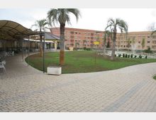 Cortile esterno con vialetto pavimentato che curva attorno a un’area verde con palme e piccole siepi. Sullo sfondo si vedono edifici a più piani in mattoni e intonaco e, a sinistra, una struttura coperta tipo pergolato con tavoli e sedie.