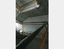 Interno di una tromba delle scale in cemento grezzo, con rampe sovrapposte e pianerottoli angolari. Ringhiere metalliche con rete e corrimano scuro in primo piano lungo il percorso.
