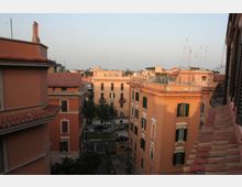 Veduta dall’alto su un quartiere urbano con palazzi residenziali color ocra e rosa, finestre con persiane verdi e tetti in tegole rosse. Tra gli edifici si intravede una strada alberata e, sullo sfondo, altri tetti con antenne sotto un cielo sereno.