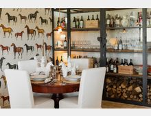 Sala da pranzo con tavolo rotondo in legno apparecchiato e sedie bianche, decorata con candelabri e centrotavola a forma di cavallo. Sullo sfondo, grande armadio vetrato con bottiglie di vino, bicchieri e legnaia in basso, e parete con carta da parati raffigurante cavalli.
