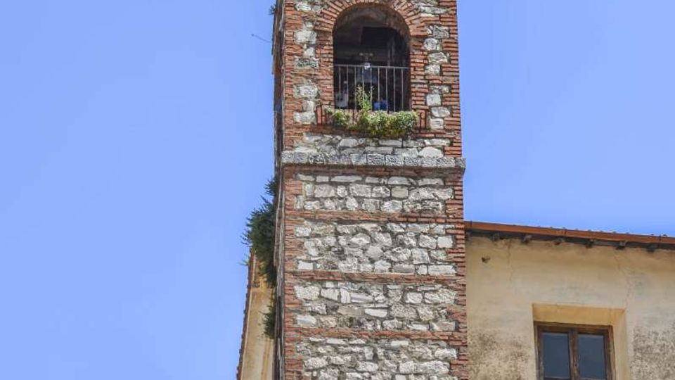 Campanile in pietra e mattoni con cuspide piramidale, caratterizzato da aperture ad arco e una cella campanaria con ringhiera. La torre è addossata a edifici in muratura in un contesto di borgo, sullo sfondo di cielo azzurro.