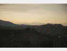 Panorama di colline ondulate con vegetazione e filari di cipressi in primo piano, con un piccolo borgo su un crinale sulla destra. Sullo sfondo si vedono montagne stratificate in lontananza sotto un cielo chiaro al tramonto.
