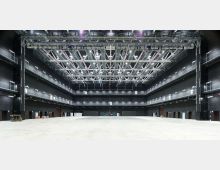 Ampio spazio interno tipo studio di produzione o palazzetto industriale, con pareti nere e passerelle perimetrali su più livelli con ringhiere metalliche. Soffitto alto con griglia di travi e tralicci illuminati, e grande pavimento libero in cemento.