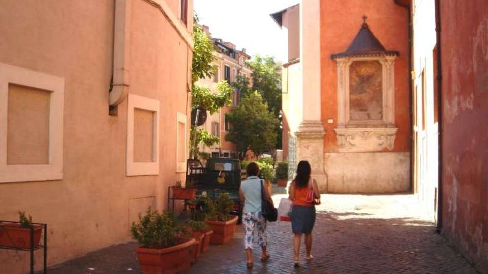 Vicolo acciottolato tra edifici dai toni ocra e rosa, con fioriere rettangolari lungo il bordo e un piccolo veicolo parcheggiato sul lato. Sulla facciata di un edificio si vede un’edicola sacra con affresco incorniciata in pietra, illuminata dalla luce del sole.