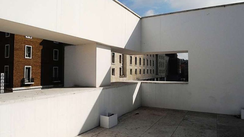 Terrazza in cemento di un edificio moderno, con alti muri bianchi e ampie aperture rettangolari che incorniciano la vista su palazzi residenziali vicini. Pavimentazione in lastre e cielo parzialmente nuvoloso sullo sfondo.