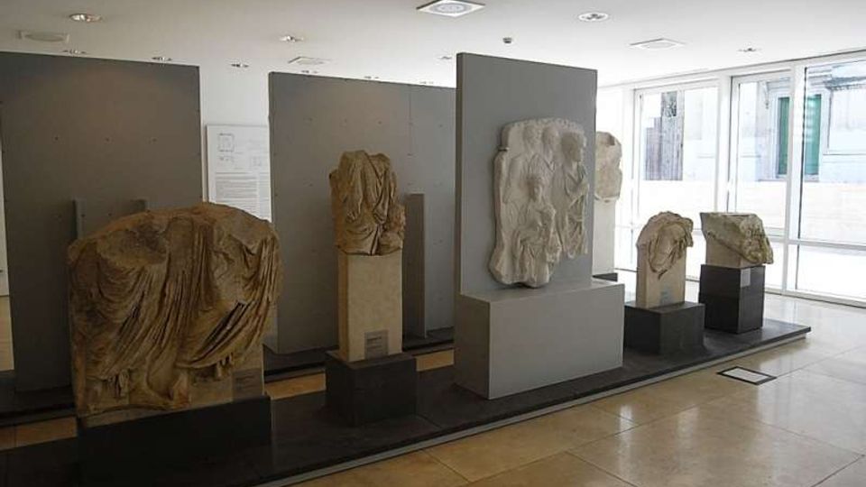Interno di una sala museale moderna con pavimento in pietra chiara, pareti espositive grigie e illuminazione a soffitto, con rilievi e frammenti scultorei in pietra su piedistalli. Grandi finestre sul lato destro lasciano entrare luce naturale e mostrano edifici all’esterno.