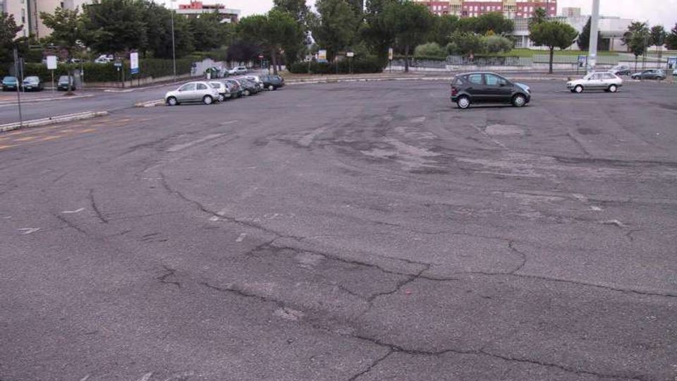 Ampio parcheggio asfaltato quasi vuoto con segnaletica a terra sbiadita e alcune auto lungo il bordo. Sullo sfondo si vedono alberi, una strada e edifici urbani in lontananza.