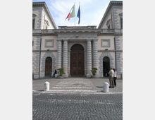 Facciata di un edificio istituzionale in stile neoclassico con grande portale centrale in legno, colonne bugnate e archi laterali. Davanti c’è una piazza lastricata in sampietrini con dissuasori e vasi, e sul tetto sventolano le bandiere italiana e dell’Unione Europea.
