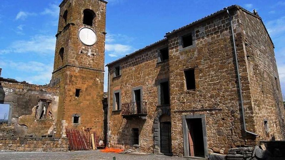 Piazza lastricata in pietra con edifici rurali in muratura di tufo, tra cui una torre campanaria con quadrante circolare e un fabbricato a due piani con piccole finestre e portali ad arco. Sullo sfondo cielo azzurro e alcune strutture parzialmente diroccate ai margini della piazza.