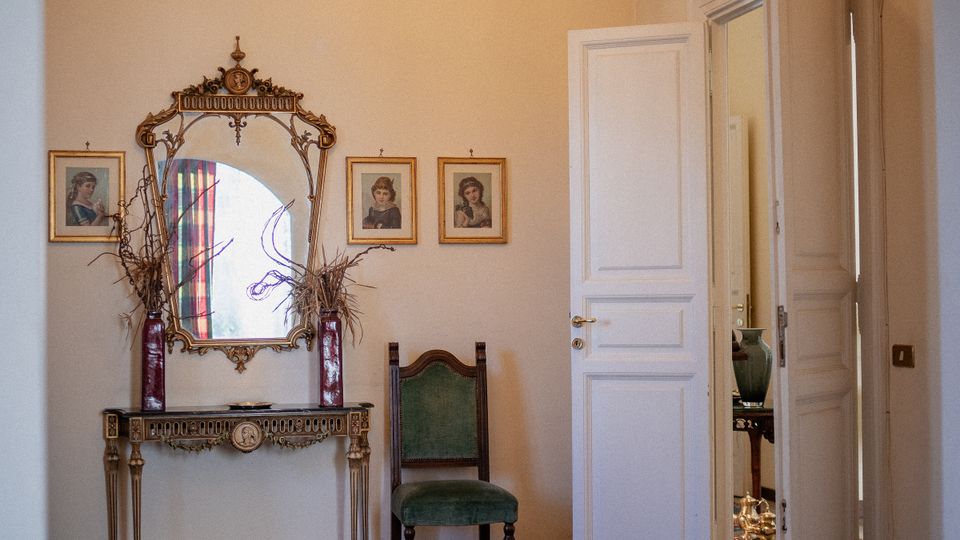 Interno di un ingresso o salotto con grande arco, pavimento in parquet e tappeto orientale; sulla parete un tavolino consolle con specchio decorato e piccoli ritratti incorniciati, con sedia verde accanto. A destra una porta bianca aperta conduce a un’altra stanza, con lampadario a cristalli sospeso in alto.