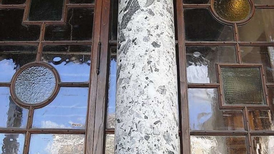 Dettaglio di una facciata con grande finestra a riquadri in legno e inserti di vetro colorato circolari e quadrati. Al centro si vede una colonna in marmo screziato su base chiara, davanti al serramento.