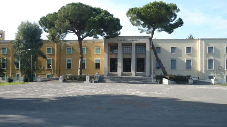 Edificio pubblico a due piani con facciata simmetrica e ingresso centrale incorniciato da colonne e una breve scalinata. Davanti si apre un ampio piazzale asfaltato con due grandi pini marittimi ai lati dell’entrata.