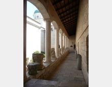 Loggiato in pietra di un edificio storico con lunga fila di colonne e archi, pavimento lastricato e tetto con travi in legno. Sul parapetto sono esposti capitelli e frammenti lapidei, con vista su un cortile e una torre in pietra sullo sfondo.