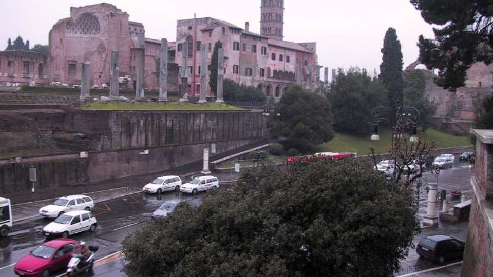 Veduta urbana con un complesso storico in mattoni e una torre sullo sfondo, affiancato da rovine con colonne e mura antiche. In primo piano si vede una strada bagnata dalla pioggia con auto in transito e aree verdi con alberi.