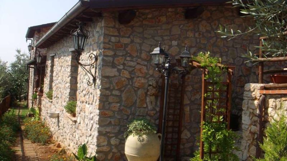 Edificio rustico in pietra con tetto sporgente e lanterne in ferro battuto fissate alla parete, lungo un vialetto in mattoni. All’esterno si vedono un grande vaso in terracotta, una struttura a graticcio con piante rampicanti e vegetazione da giardino ai lati.