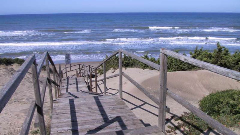 Passerella e scala in legno con corrimano che scende tra dune di sabbia e vegetazione verso una spiaggia. Sullo sfondo si vede il mare aperto con onde e l’orizzonte sotto un cielo sereno.