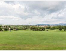 Ampio green di un campo da golf con bandierina al centro, circondato da prato rasato e alberi. Sullo sfondo si vedono colline e una valle con edifici sparsi sotto un cielo nuvoloso.