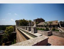 Terrazza panoramica in pietra su bastioni murati, con parapetto scandito da pilastri e pavimentazione in mattoni. Sullo sfondo si vedono edifici storici con tetti in coppi, palme e alberi, sotto un cielo sereno.
