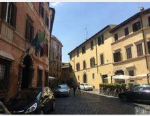 Strada urbana stretta con pavimentazione in ciottoli, fiancheggiata da edifici storici a più piani con facciate in intonaco e finestre con persiane. Auto parcheggiate lungo i lati e, sul lato destro, un’area con tavolini e ombrelloni davanti a un edificio.