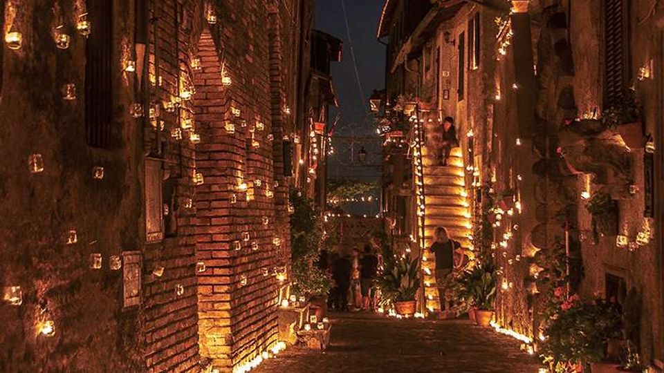 Vicolo stretto in pietra tra edifici in mattoni e intonaco, illuminato di notte da numerose candele in piccoli lumini appesi alle pareti e disposti lungo il percorso. La strada in lieve discesa è fiancheggiata da finestre, porte e vasi con piante, creando un corridoio luminoso tra le facciate.