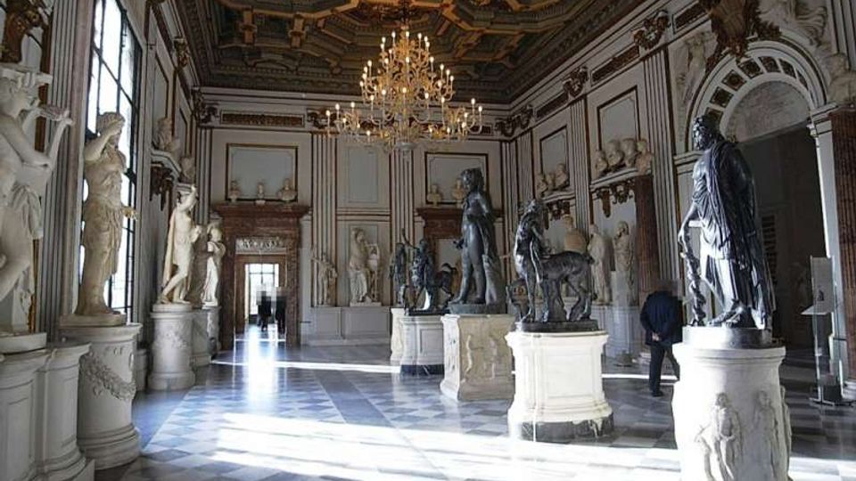 Ampia sala museale in stile neoclassico con pavimento a scacchi in marmo, grandi finestre laterali e soffitto a cassettoni decorato con lampadari. Numerose statue su piedistalli e nicchie lungo le pareti ornano lo spazio, con un passaggio in fondo alla sala.