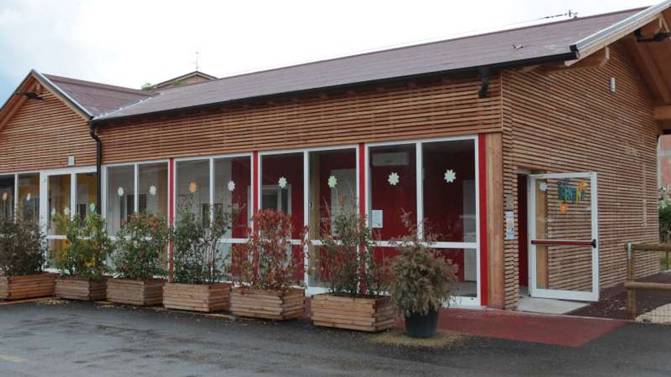 Edificio basso in legno con tetto a falde e lunga facciata vetrata incorniciata di rosso, con finestre decorate e un ingresso laterale. Davanti si trovano fioriere rettangolari con arbusti lungo un piazzale asfaltato.
