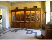 Interno di sala museale con pareti gialle e pavimento in marmo a motivi geometrici, dominata da una grande vetrina in legno illuminata che espone vasi e ceramiche. A destra è visibile un’altra teca in vetro, mentre a sinistra si apre un passaggio verso un ambiente adiacente.