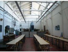 Ampio laboratorio/aula tecnica con lunghi tavoli e sedie disposti in file, pavimento rosso e attrezzature industriali lungo le pareti. Soffitto alto con capriate bianche e lucernari longitudinali che illuminano l’ambiente.
