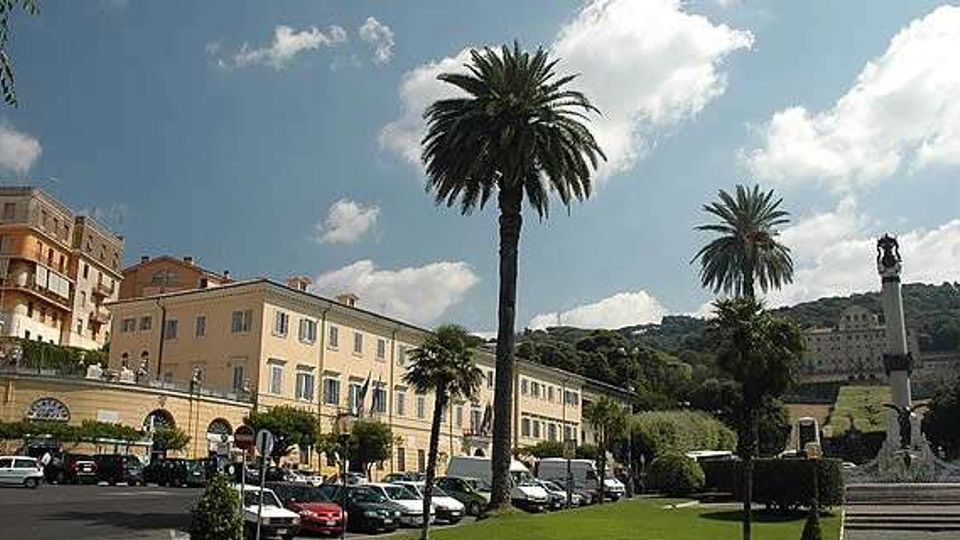Ampia piazza urbana con parcheggio e prati curati, fiancheggiata da alti palmeti e da edifici storici color pastello con archi al piano terra. Sullo sfondo si vede una collina verde e, a destra, una fontana monumentale con statua sotto un cielo parzialmente nuvoloso.