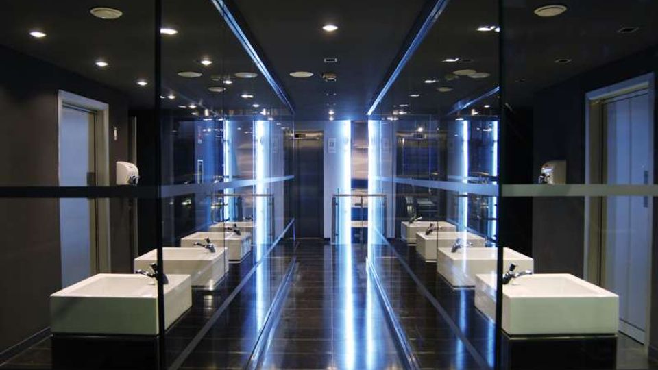 Bagno moderno con pareti specchiate e illuminazione a LED verticali lungo la parete centrale, che riflettono su pavimento scuro lucido e soffitto con faretti incassati. Ai lati ci sono lavabi bianchi squadrati con rubinetti cromati e porte su entrambe le estremità del locale.