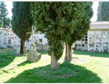 Area di cimitero con prato e grandi cipressi in primo piano, con un monumento a croce su una base in pietra. Sullo sfondo si vedono edifici con loculi disposti in file e decorati con fiori e lapidi.